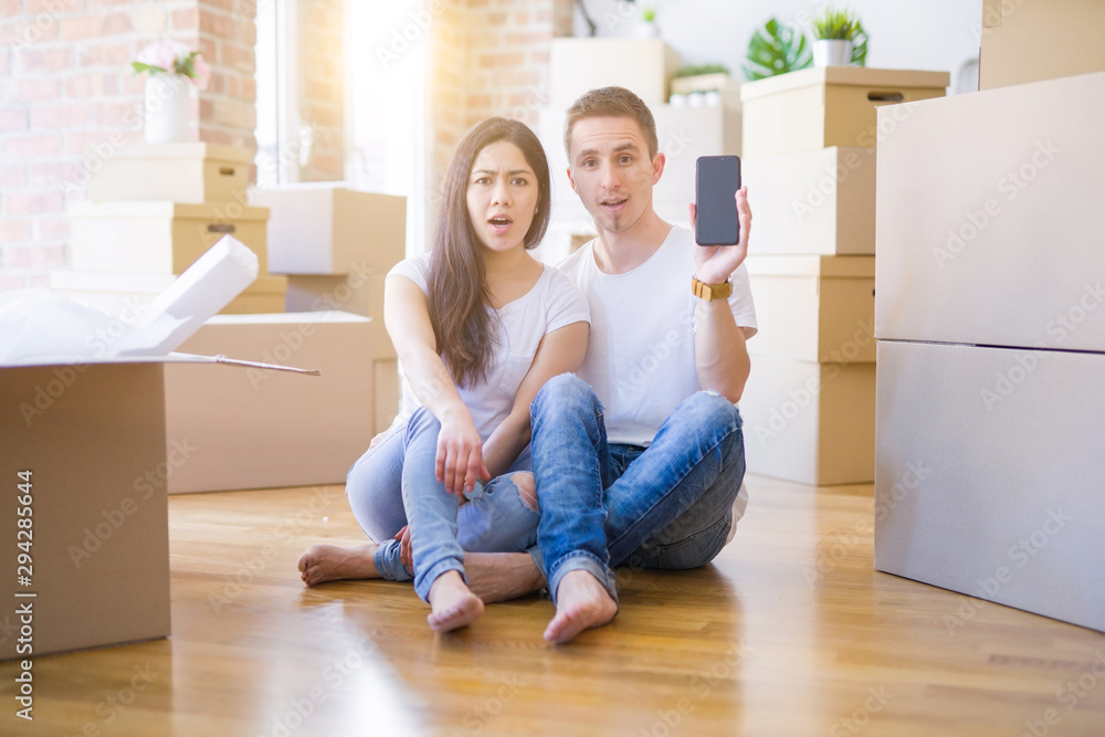 Beautiful couple sitting on the floor holding smartphone at new home around cardboard boxes scared in shock with a surprise face, afraid and excited with fear expression Beautiful couple sitting on the floor holding smartphone at new home around cardboard boxes scared in shock with a surprise face, afraid and excited with fear expression