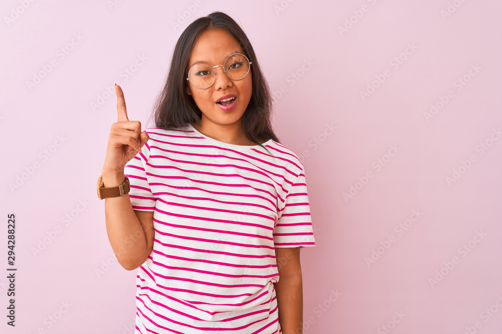 Young chinese woman wearing striped t-shirt and glasses over isolated pink background pointing finger up with successful idea. Exited and happy. Number one. Young chinese woman wearing striped t-shirt and glasses over isolated pink background pointing finger up with successful idea. Exited and happy. Number one.