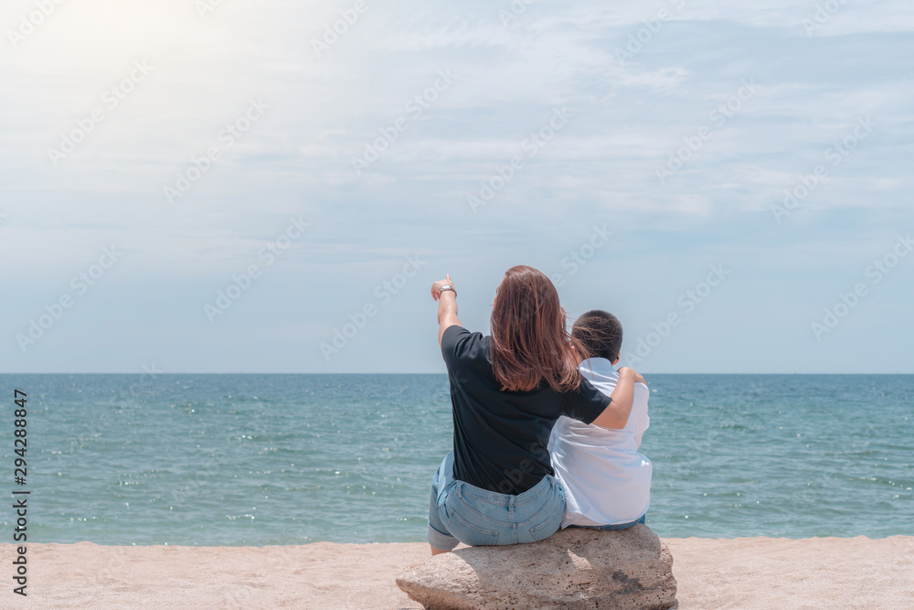Rear view of mother hugs a son by the shoulders sitting on the beach and looking out to sea