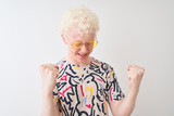 Young albino blond man wearing colorful t-shirt and sunglasses over isolated red background very happy and excited doing winner gesture with arms raised, smiling and screaming for success