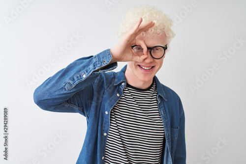 Young albino blond man wearing denim shirt and glasses over isolated white background doing ok gesture with hand smiling, eye looking through fingers with happy face.