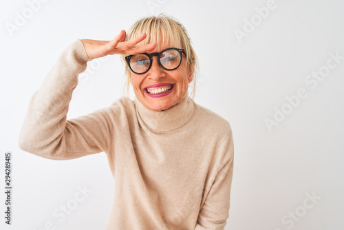 Middle age woman wearing turtleneck sweater and glasses over isolated white background very happy and smiling looking far away with hand over head. Searching concept.