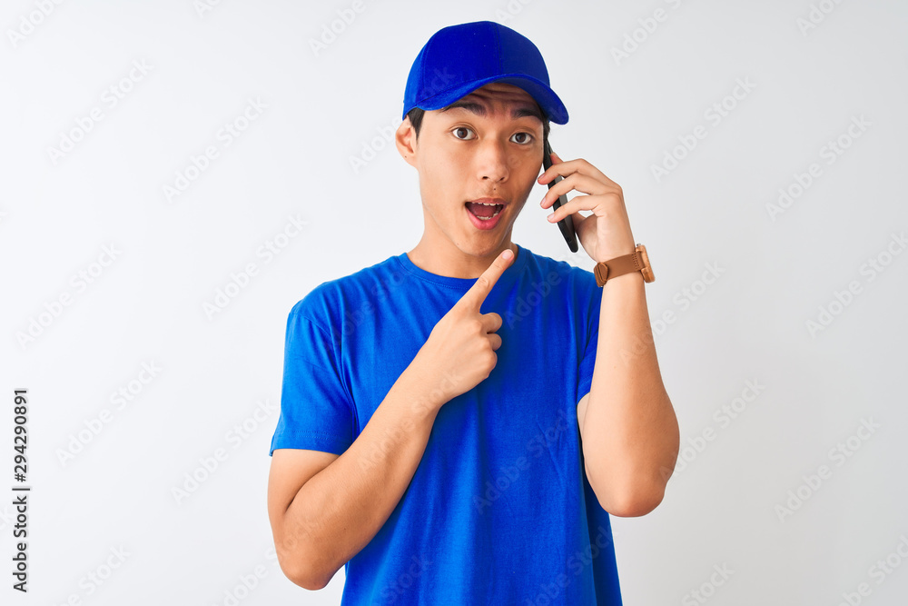 Chinese deliveryman wearing cap talking on the smartphone over isolated white background very happy pointing with hand and finger