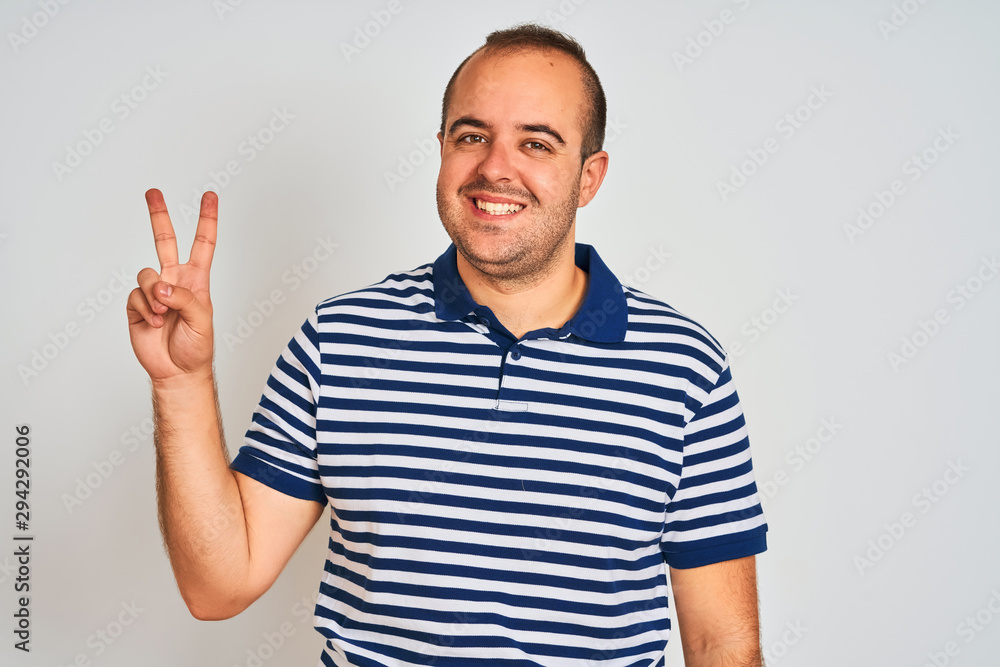 Fototapeta premium Young man wearing casual striped polo standing over isolated white background smiling looking to the camera showing fingers doing victory sign. Number two.