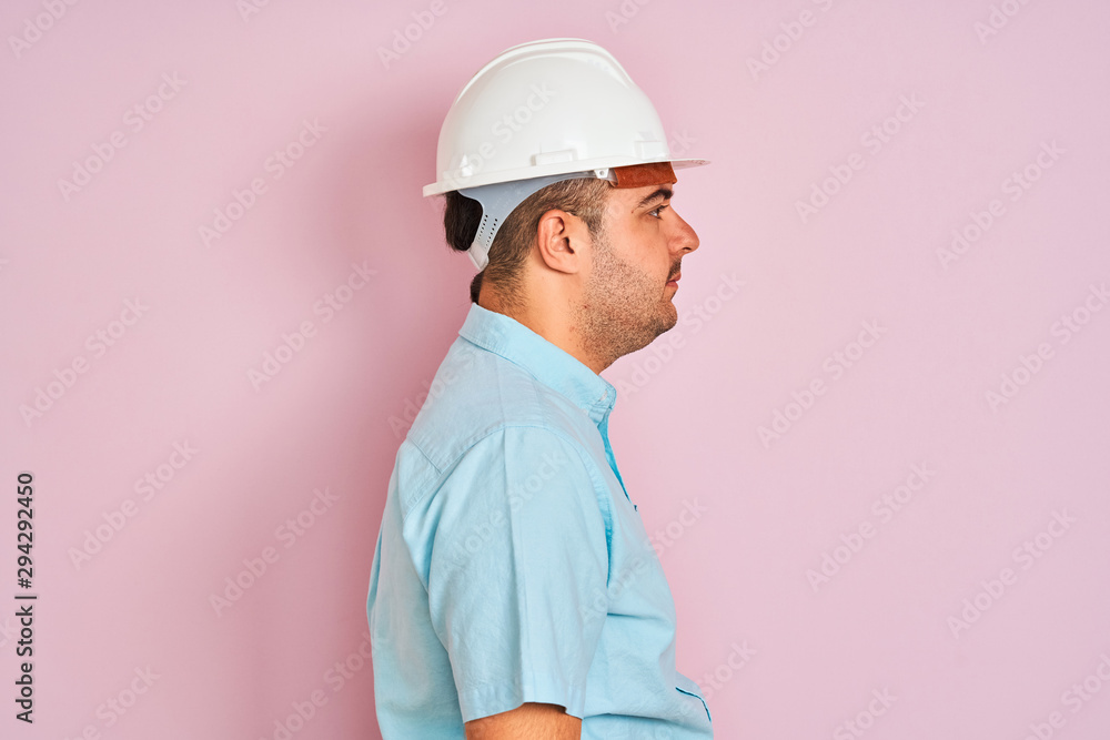 Young architect man wearing security helmet standing over isolated pink background looking to side, relax profile pose with natural face with confident smile.
