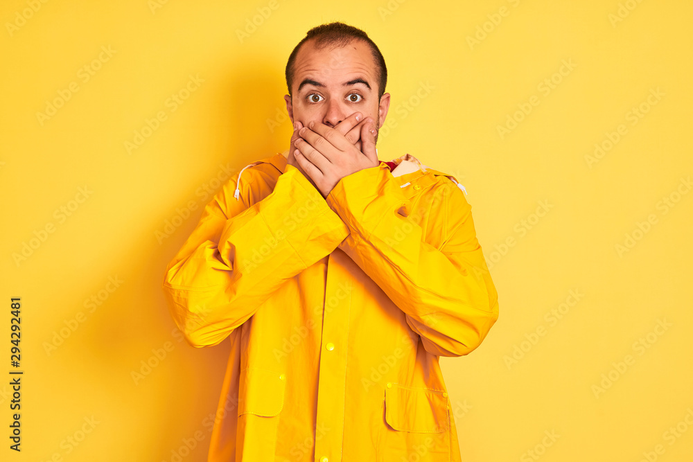 Young man wearing rain coat standing over isolated yellow background shocked covering mouth with hands for mistake. Secret concept.