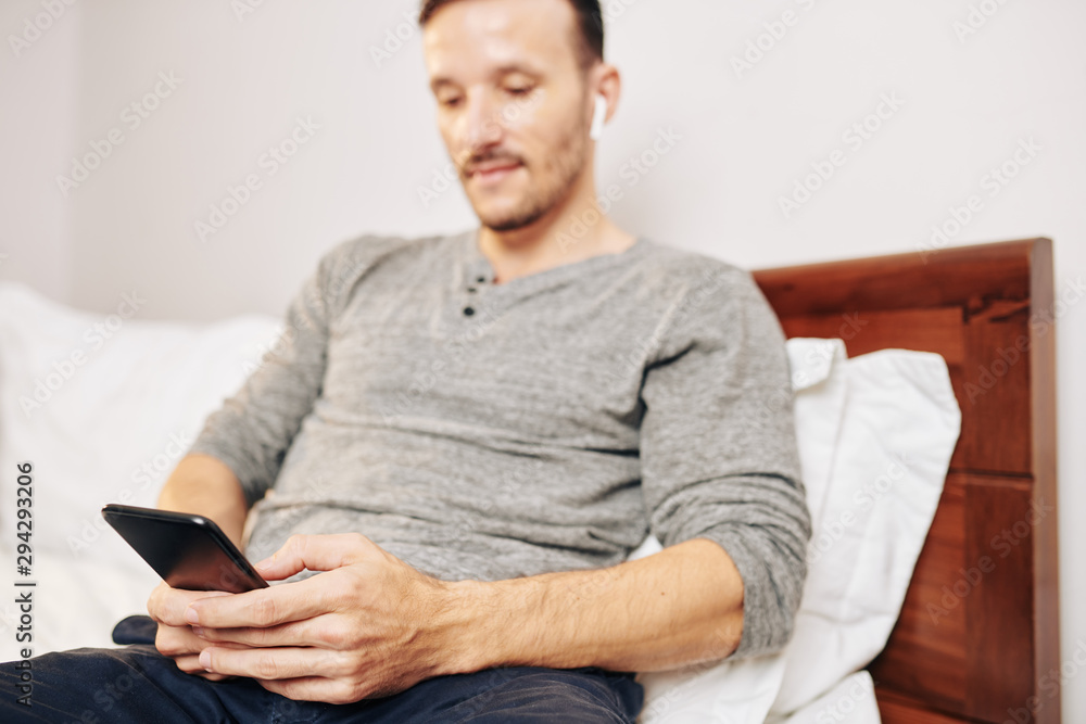 Young man sitting in bedroom, texting friends and checking social media, focus on smartphone Young man sitting in bedroom, texting friends and checking social media, focus on smartphone