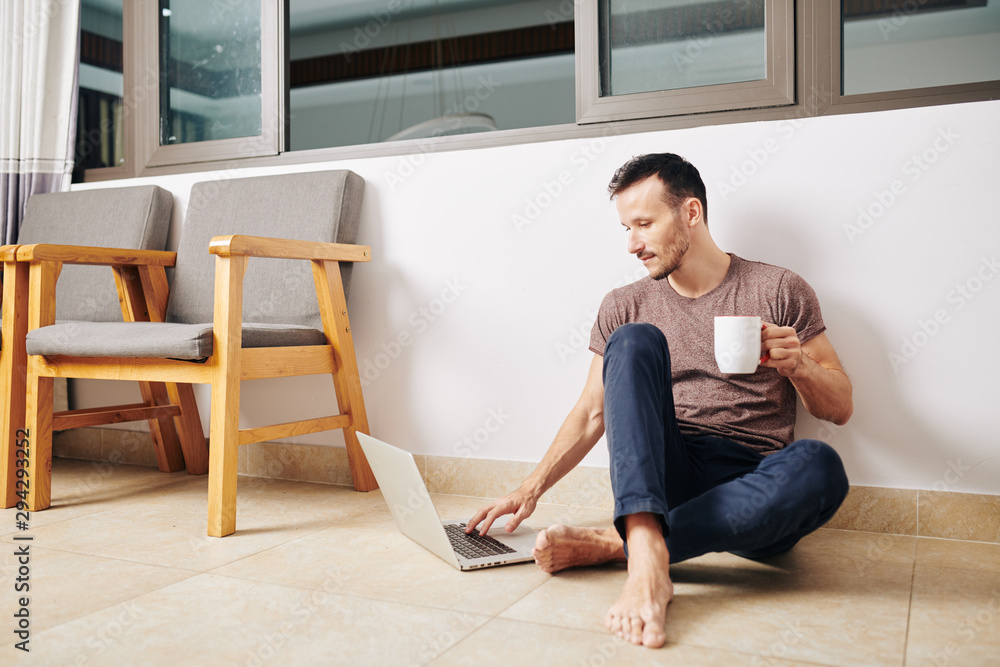 Handsome young barefoot man sitting on the floor in his room, drinking coffee and checking e-mails