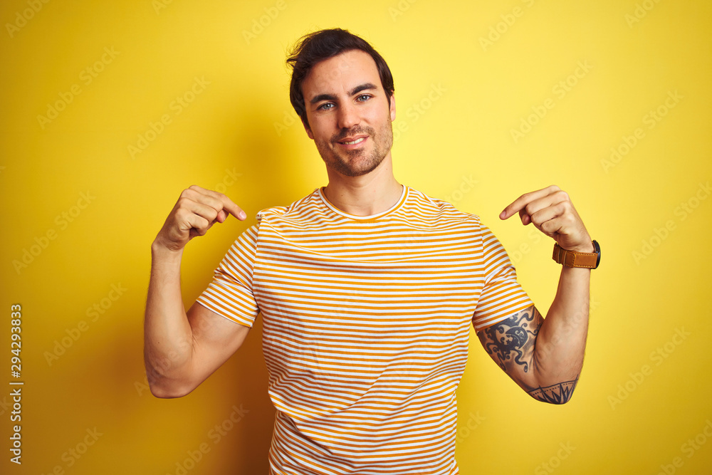 Young handsome man with tattoo wearing striped t-shirt over isolated yellow background looking confident with smile on face, pointing oneself with fingers proud and happy. Young handsome man with tattoo wearing striped t-shirt over isolated yellow background looking confident with smile on face, pointing oneself with fingers proud and happy.