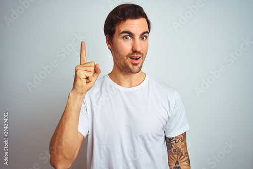 Young handsome man with tattoo wearing casual t-shirt over isolated white background pointing finger up with successful idea. Exited and happy. Number one.