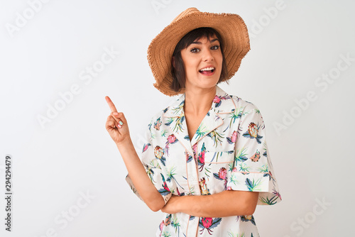 Beautiful woman on vacation wearing summer shirt and hat over isolated white background with a big smile on face, pointing with hand and finger to the side looking at the camera.