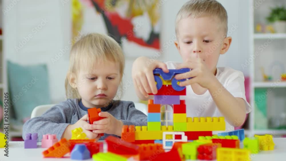 Two little boys sitting at table in kindergarten and building toy house together while playing with construction set Two little boys sitting at table in kindergarten and building toy house together while playing with construction set