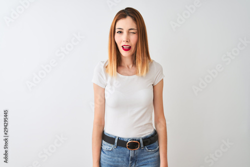 Beautiful redhead woman wearing casual t-shirt standing over isolated white background winking looking at the camera with sexy expression, cheerful and happy face.