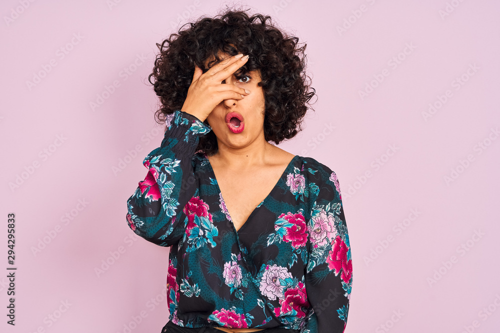 Young arab woman with curly hair wearing floral dress over isolated pink background peeking in shock covering face and eyes with hand, looking through fingers with embarrassed expression. Young arab woman with curly hair wearing floral dress over isolated pink background peeking in shock covering face and eyes with hand, looking through fingers with embarrassed expression.