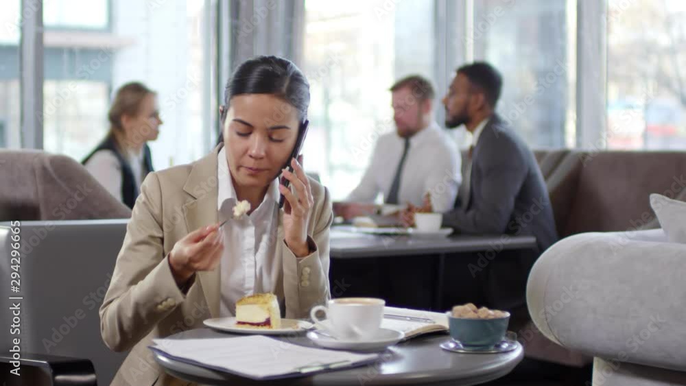 Young asian businesswoman sitting at table in coffeeshop, eating sweet cake and talking on mobile phone while having coffee break Young asian businesswoman sitting at table in coffeeshop, eating sweet cake and talking on mobile phone while having coffee break