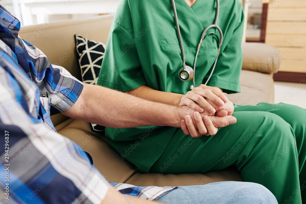 Close-up image of nurse in green scrubs holding hand of elderly patient when visiting him at home and talking about recovery process Close-up image of nurse in green scrubs holding hand of elderly patient when visiting him at home and talking about recovery process