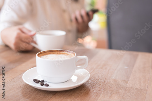 Hot coffee in the white cup and coffee beans in a sack on wooden table background with blurry woman holding coffee cup.Cafe drinking menu latte art coffee at the restaurant with copy space.