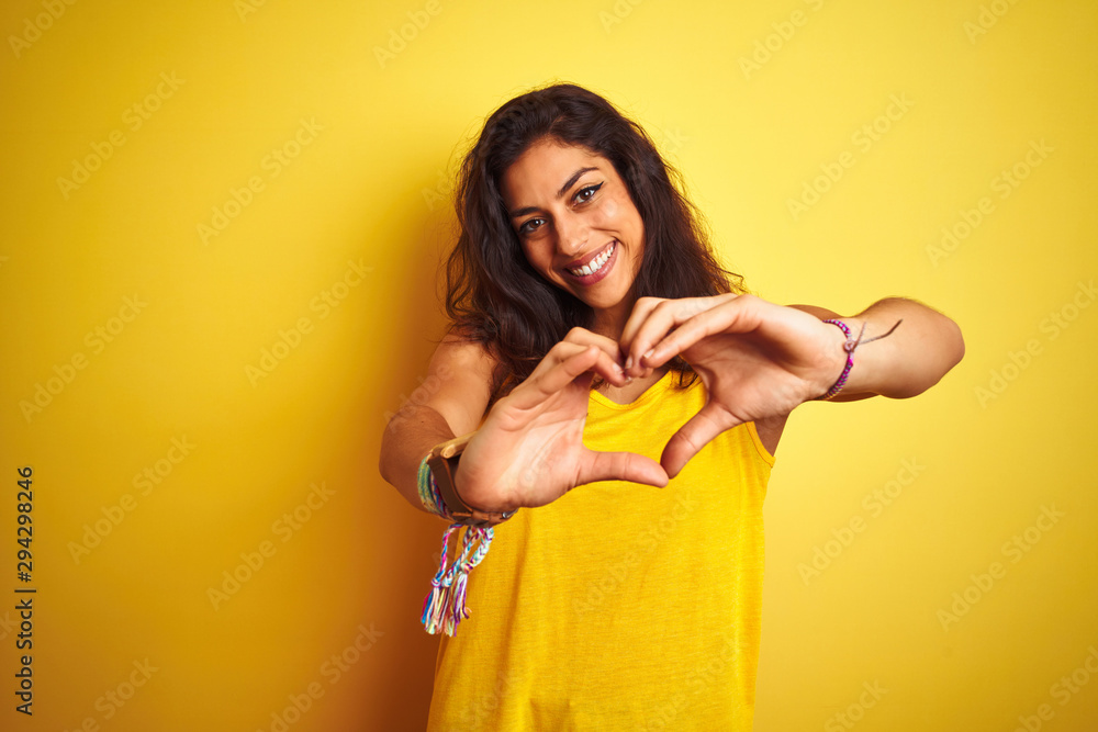 Young beautiful woman wearing t-shirt standing over isolated yellow background smiling in love showing heart symbol and shape with hands. Romantic concept. Young beautiful woman wearing t-shirt standing over isolated yellow background smiling in love showing heart symbol and shape with hands. Romantic concept.