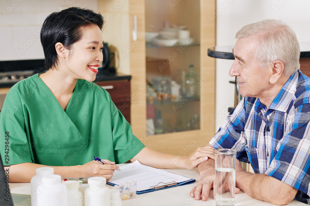 Obraz premium Smiling Vietnamese nurse touching hand of senior patient and filling his medical card when visiting him at home