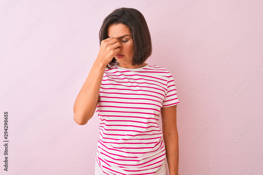 Young beautiful woman wearing striped t-shirt standing over isolated pink background tired rubbing nose and eyes feeling fatigue and headache. Stress and frustration concept. Young beautiful woman wearing striped t-shirt standing over isolated pink background tired rubbing nose and eyes feeling fatigue and headache. Stress and frustration concept.