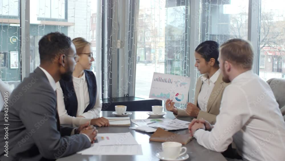 Zoom out of young asian businesswoman holding financial report and discussing diagrams with multi-ethnic colleagues during coffee break in cafe Zoom out of young asian businesswoman holding financial report and discussing diagrams with multi-ethnic colleagues during coffee break in cafe
