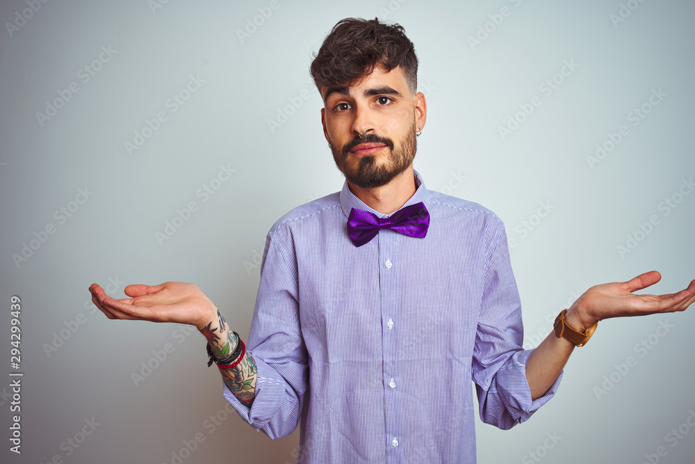 Young man with tattoo wearing purple shirt and bow tie over isolated white background clueless and confused expression with arms and hands raised. Doubt concept. Stock Photo