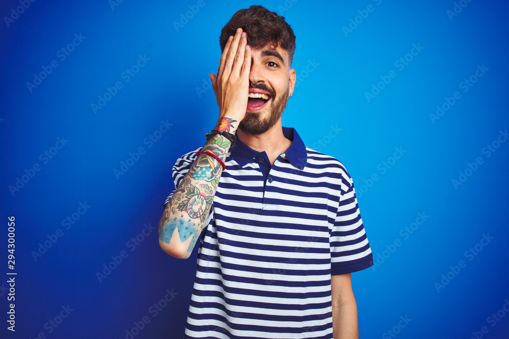 Young man with tattoo wearing striped polo standing over isolated blue background covering one eye with hand, confident smile on face and surprise emotion. Young man with tattoo wearing striped polo standing over isolated blue background covering one eye with hand, confident smile on face and surprise emotion.