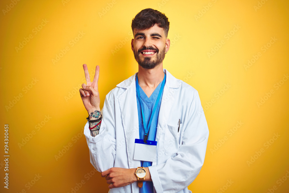Young doctor man with tattoo wearing id card standing over isolated yellow background smiling with happy face winking at the camera doing victory sign. Number two. Young doctor man with tattoo wearing id card standing over isolated yellow background smiling with happy face winking at the camera doing victory sign. Number two.