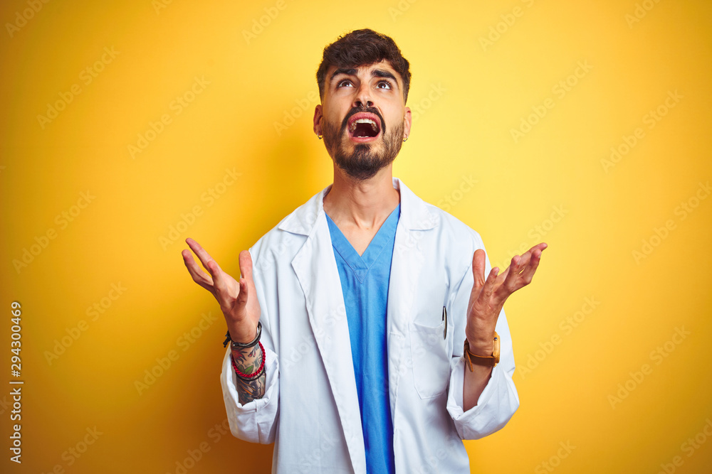 Young doctor man with tattoo standing over isolated yellow background crazy and mad shouting and yelling with aggressive expression and arms raised. Frustration concept. Young doctor man with tattoo standing over isolated yellow background crazy and mad shouting and yelling with aggressive expression and arms raised. Frustration concept.