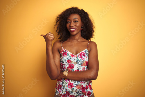 African american woman wearing floral summer t-shirt over isolated yellow background smiling with happy face looking and pointing to the side with thumb up.