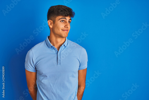 Young indian man wearing casual polo standing over isolated blue background smiling looking to the side and staring away thinking.