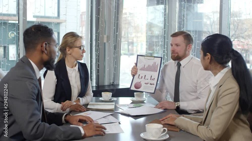 Zoom in of multi ethnic business team sitting at cafe table beside the window and discussing financial report during coffee break: Caucasian man explaining financial diagrams on clipboard photo