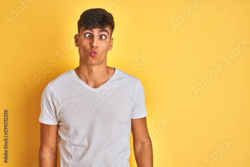Young indian man wearing white t-shirt standing over isolated yellow background making fish face with lips, crazy and comical gesture. Funny expression.