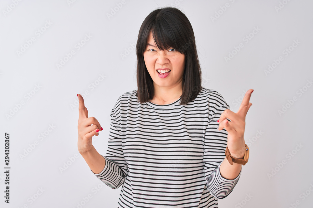 Young beautiful chinese woman wearing black striped t-shirt over isolated white background shouting with crazy expression doing rock symbol with hands up. Music star. Heavy concept. Young beautiful chinese woman wearing black striped t-shirt over isolated white background shouting with crazy expression doing rock symbol with hands up. Music star. Heavy concept.