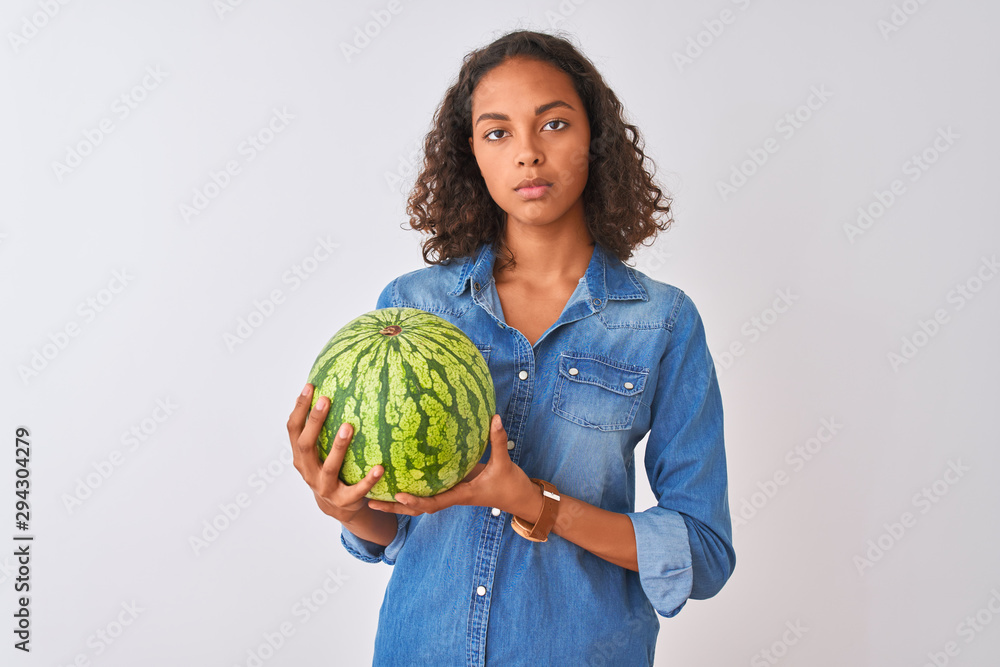 Young brazilian woman holding watermelon standing over isolated white background with a confident expression on smart face thinking serious Young brazilian woman holding watermelon standing over isolated white background with a confident expression on smart face thinking serious