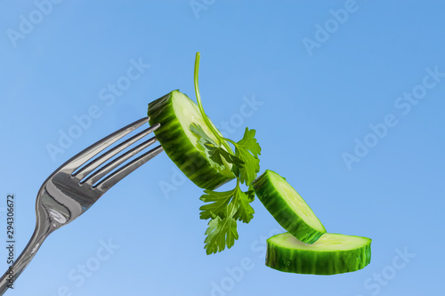 Fotografie A slice of green cucumber, impaled on a metal fork on a blue background, close-u