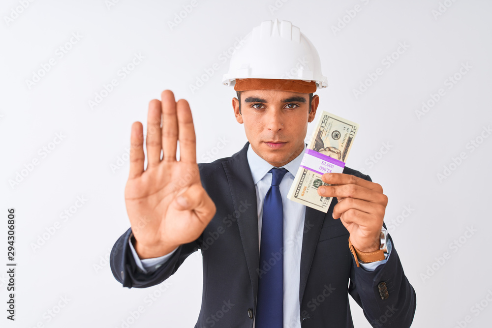 Young handsome architect man wearing helmet holding dollars over isolated white background with open hand doing stop sign with serious and confident expression, defense gesture Young handsome architect man wearing helmet holding dollars over isolated white background with open hand doing stop sign with serious and confident expression, defense gesture