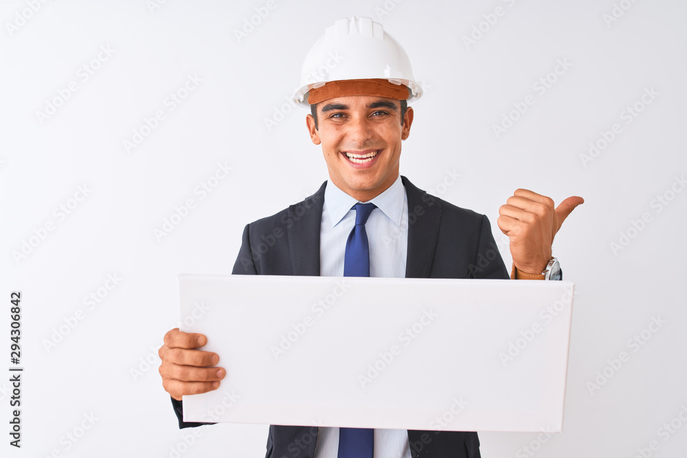 Young handsome architect man wearing helmet holding banner over isolated white background pointing and showing with thumb up to the side with happy face smiling Young handsome architect man wearing helmet holding banner over isolated white background pointing and showing with thumb up to the side with happy face smiling