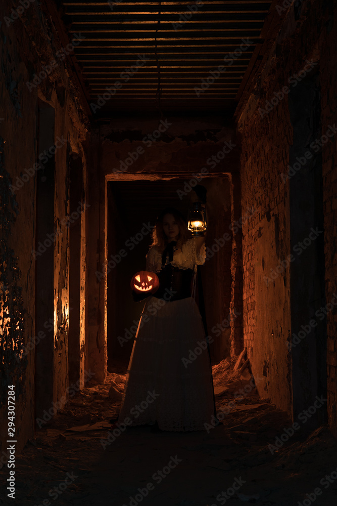 Girl in vintage dress standing in dark corridor holding halloween pumpkin glowing from inside and oil lantern