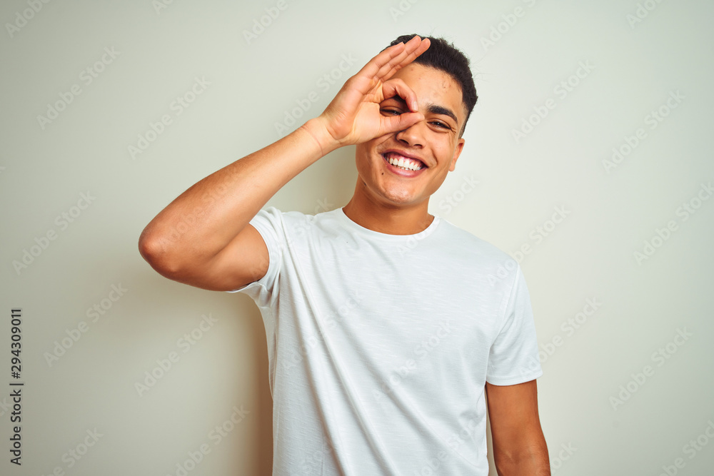 Young brazilian man wearing t-shirt standing over isolated white background doing ok gesture with hand smiling, eye looking through fingers with happy face. Young brazilian man wearing t-shirt standing over isolated white background doing ok gesture with hand smiling, eye looking through fingers with happy face.