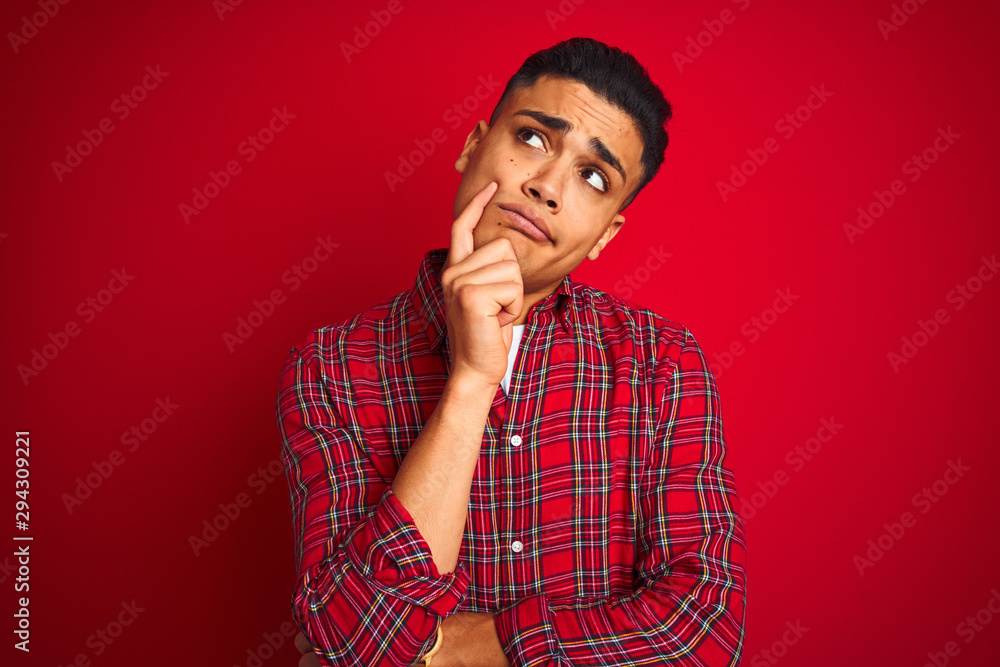 Young brazilian man wearing shirt standing over isolated red background with hand on chin thinking about question, pensive expression. Smiling with thoughtful face. Doubt concept. Young brazilian man wearing shirt standing over isolated red background with hand on chin thinking about question, pensive expression. Smiling with thoughtful face. Doubt concept.