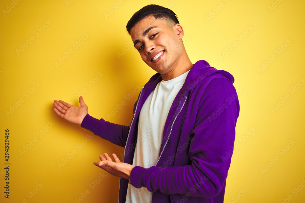 Young brazilian man wearing purple sweatshirt standing over isolated yellow background Inviting to enter smiling natural with open hand Young brazilian man wearing purple sweatshirt standing over isolated yellow background Inviting to enter smiling natural with open hand