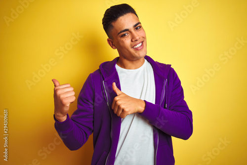 Young brazilian man wearing purple sweatshirt standing over isolated yellow background Pointing to the back behind with hand and thumbs up, smiling confident photo