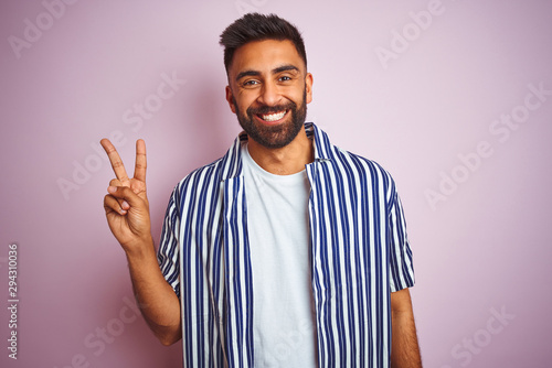 Young handsome indian man wearing summer striped shirt over isolated pink background smiling with happy face winking at the camera doing victory sign. Number two.