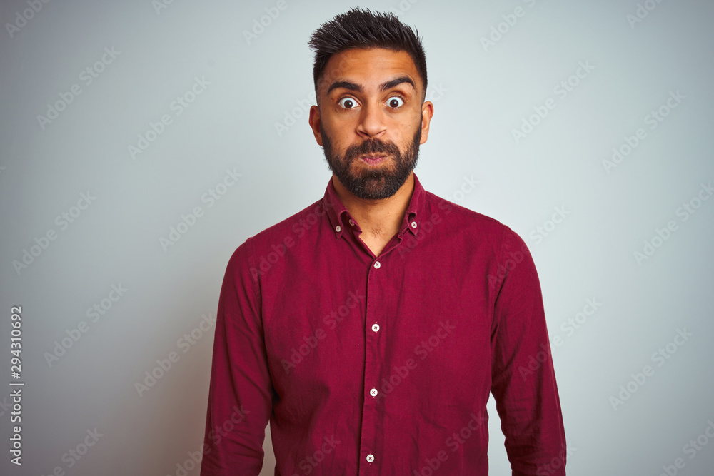 Obraz premium Young indian man wearing red elegant shirt standing over isolated grey background puffing cheeks with funny face. Mouth inflated with air, crazy expression.