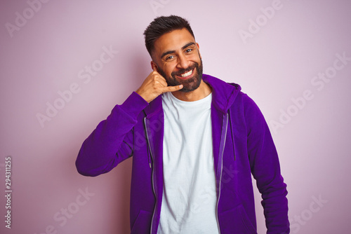 Young indian man wearing purple sweatshirt standing over isolated pink background smiling doing phone gesture with hand and fingers like talking on the telephone. Communicating concepts. © Krakenimages.com