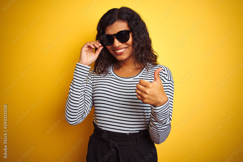 Transsexual transgender woman wearing sunglasses over isolated yellow background doing happy thumbs up gesture with hand. Approving expression looking at the camera with showing success.
