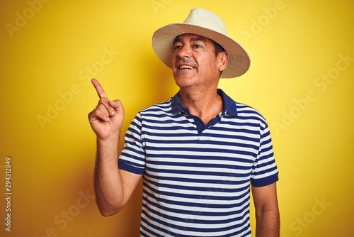 Handsome middle age man wearing striped polo and hat over isolated yellow background with a big smile on face, pointing with hand and finger to the side looking at the camera.