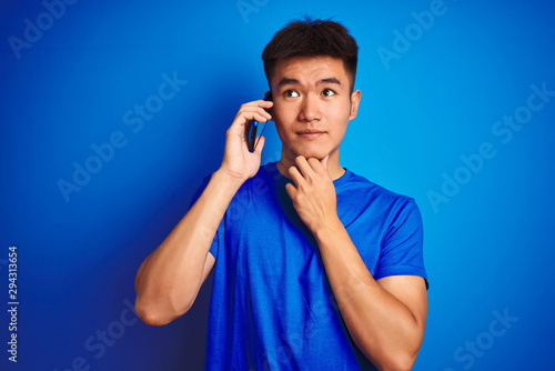 Young asian chinese man talking on the smartphone standing over isolated blue background serious face thinking about question, very confused idea
