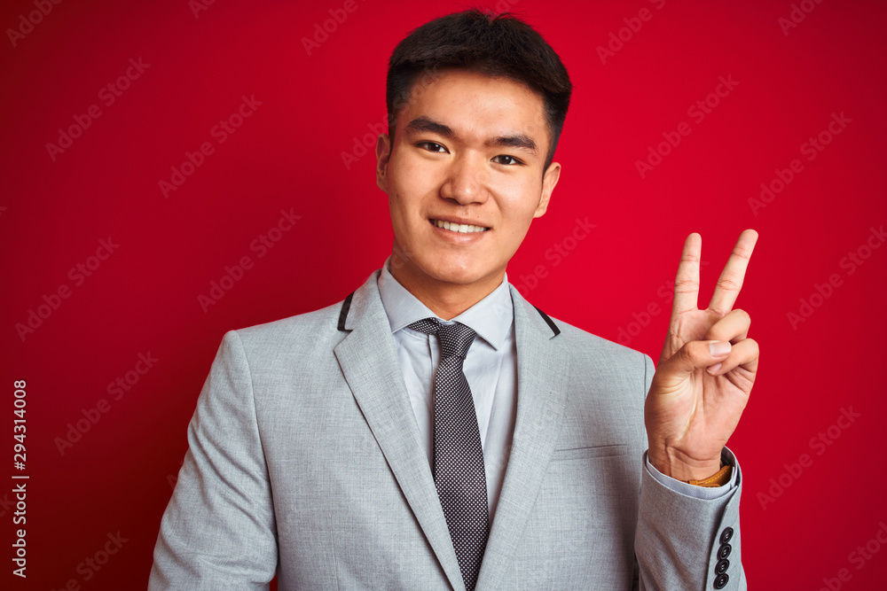 Asian chinese businessman wearing grey jacket and tie standing over isolated red background smiling with happy face winking at the camera doing victory sign. Number two. Asian chinese businessman wearing grey jacket and tie standing over isolated red background smiling with happy face winking at the camera doing victory sign. Number two.